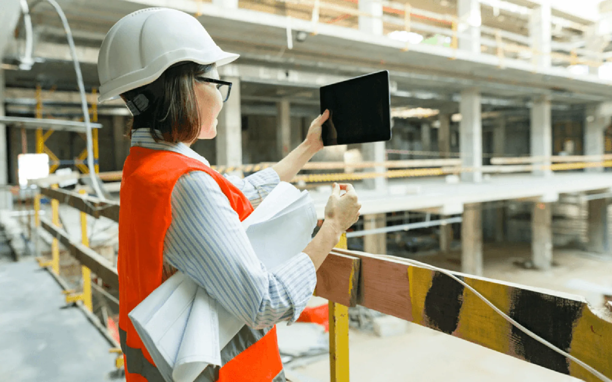 Eine Bauleiterin trägt einen Schutzhelm und eine Sicherheitsweste, während sie mit einem Tablet und Bauplänen eine Baustelle überwacht.