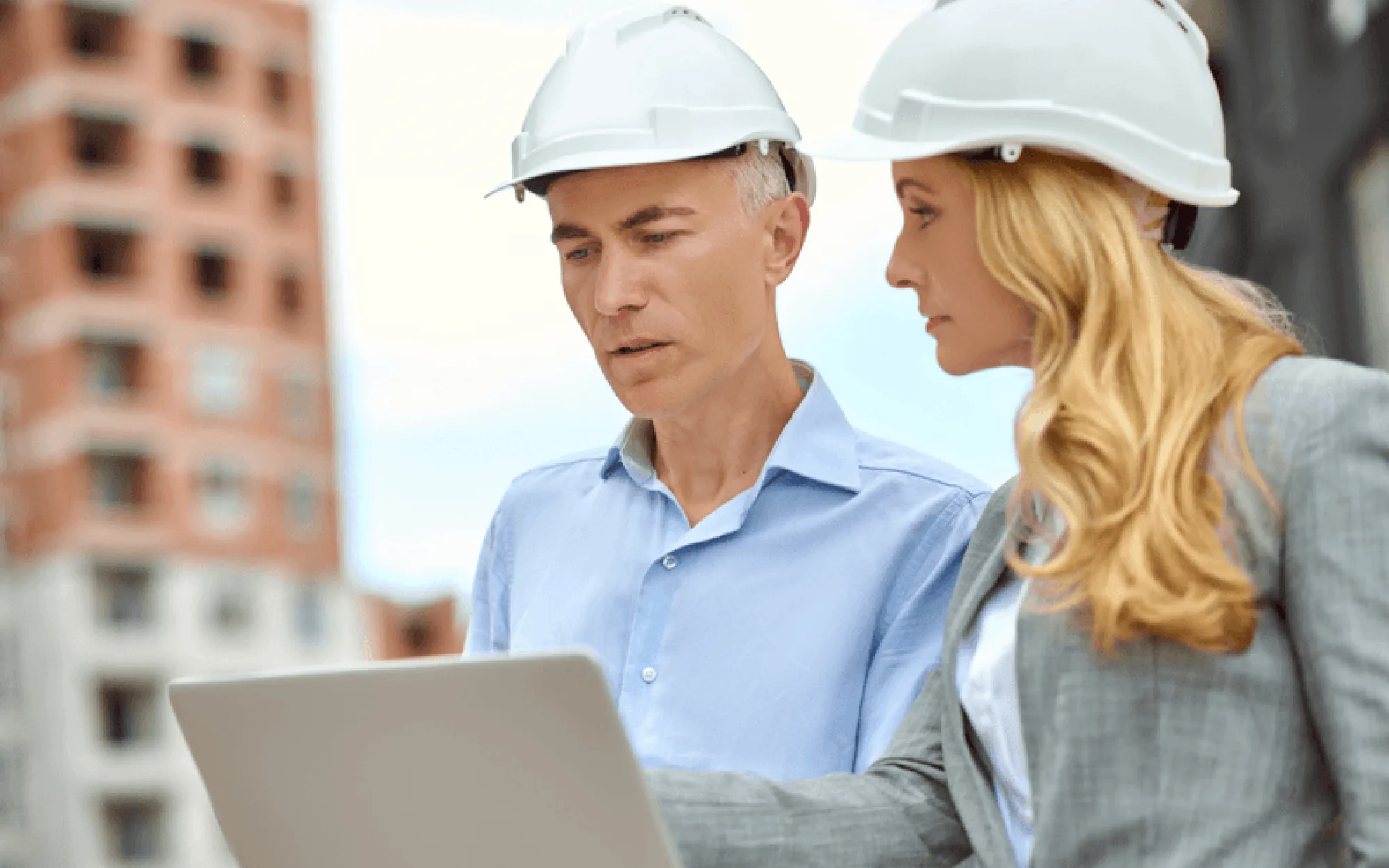 Zwei Architekten mit weißen Schutzhelmen besprechen sich auf einer Baustelle, während sie gemeinsam auf einen Laptop schauen.