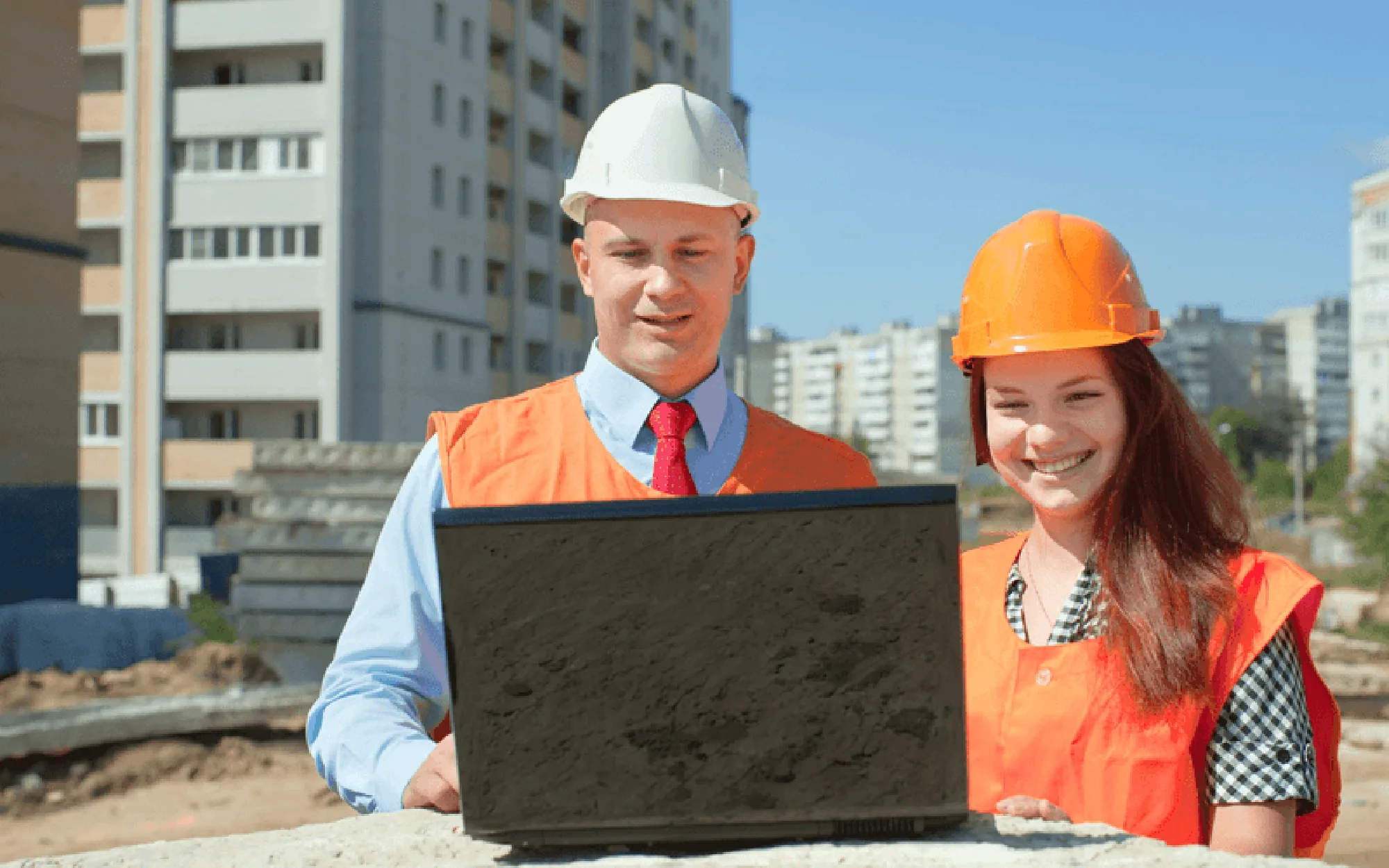 Ein Bauleiter und eine Bauleiterin tragen Schutzhelme und orangefarbene Westen, während sie auf einer Baustelle gemeinsam an einem Laptop arbeiten.