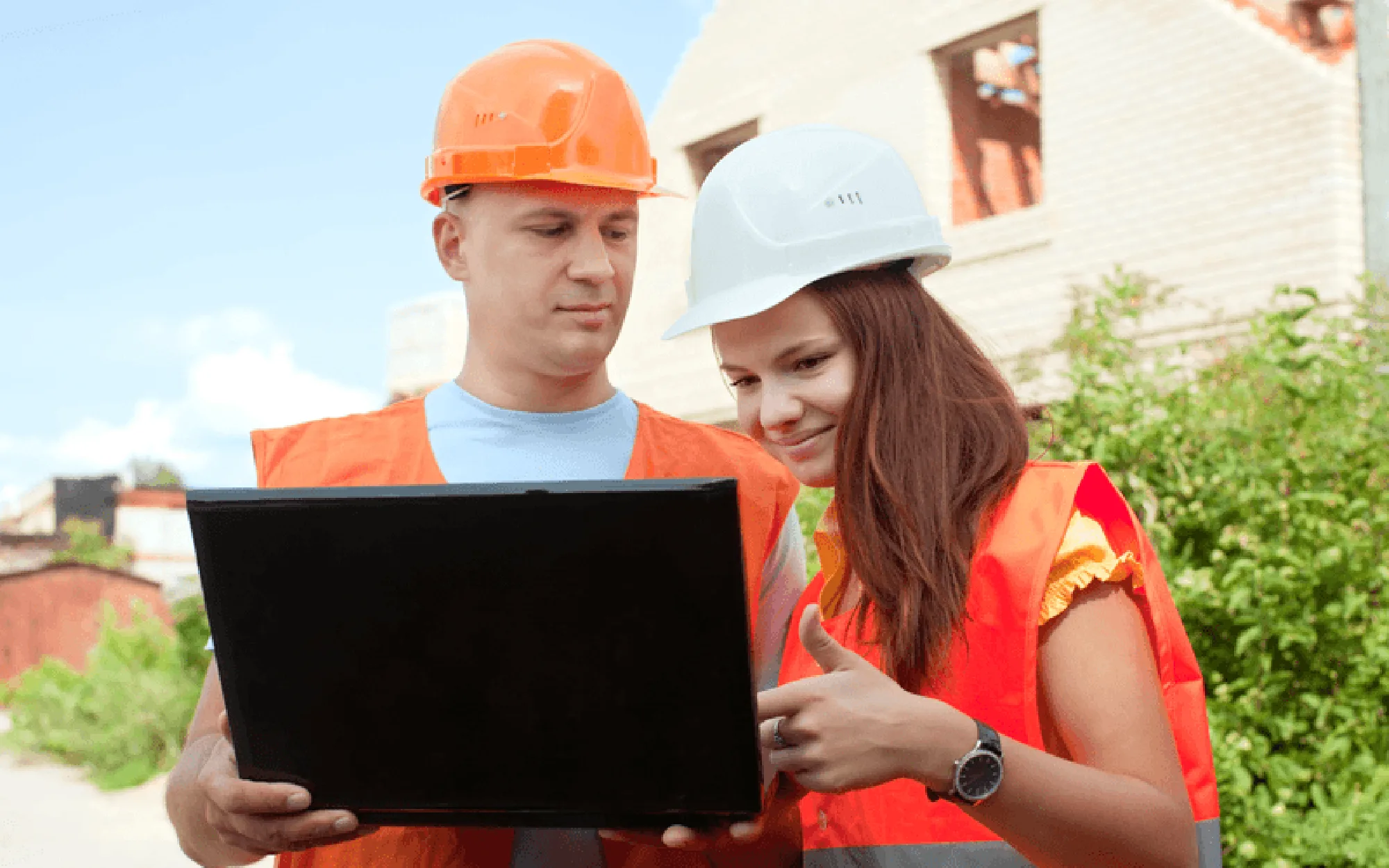 Two site managers wearing hard hats work together on a laptop on an outdoor construction site.